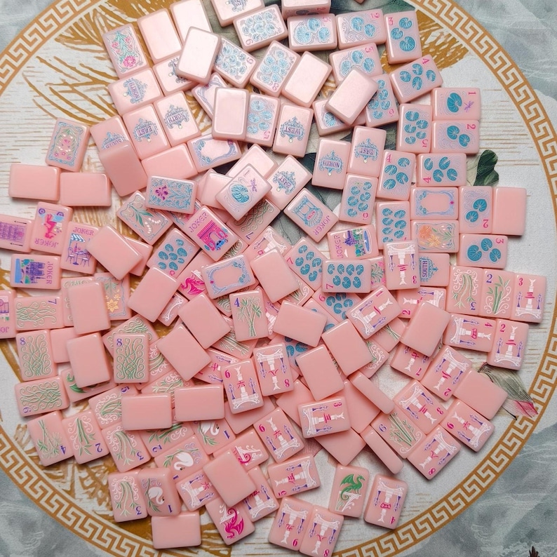 May include: A collection of rectangular pink mahjong tiles scattered across a surface. Each tile features engraved designs in blue, green, and pink, including numbers, directional symbols, floral patterns, and joker icons with intricate line work.