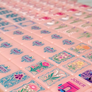 May include: A set of light pink Mahjong tiles arranged in rows, featuring intricate blue and multi-colored illustrations of flowers, directional markers like East, West, and South, and tiles labeled Joker with detailed architectural scenes.