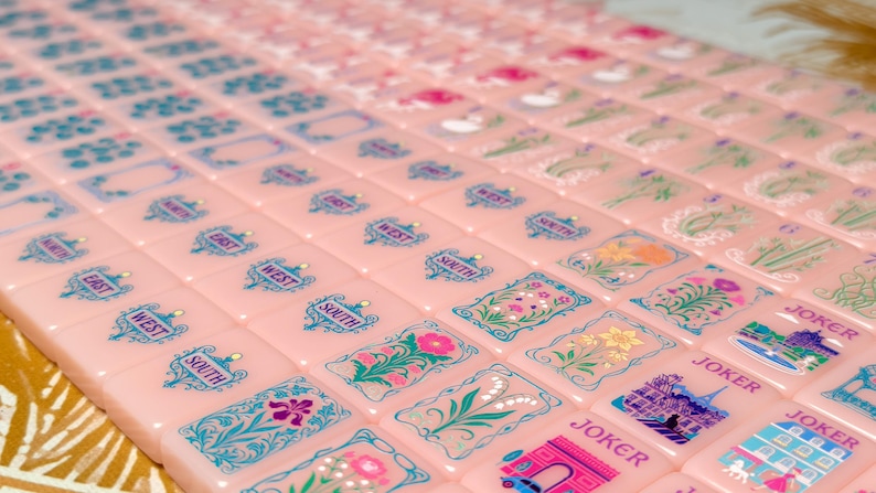 May include: A set of light pink Mahjong tiles arranged in rows, featuring intricate blue and multi-colored illustrations of flowers, directional markers like East, West, and South, and tiles labeled Joker with detailed architectural scenes.