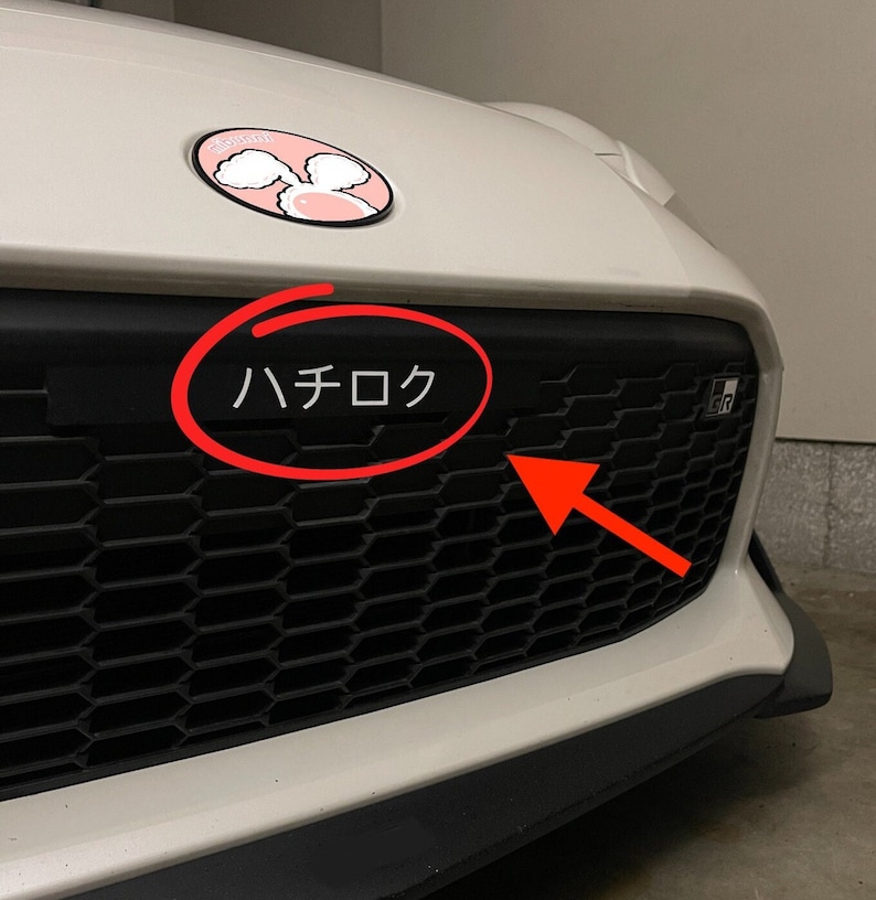 May include: White car with a black grill and a black plate that says "ハチロク" in Japanese characters. The car has a pink cartoon sheep sticker on the hood.