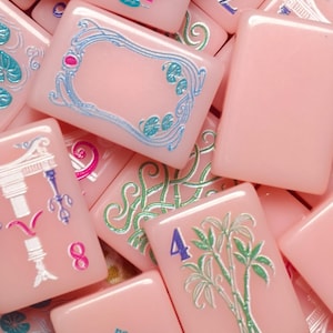 May include: A collection of rectangular mahjong tiles in a soft pink hue, featuring engraved designs in metallic blue, green, and silver. The tiles display various symbols, including the word JOKER, the word NORTH, a bamboo plant, and numbers.