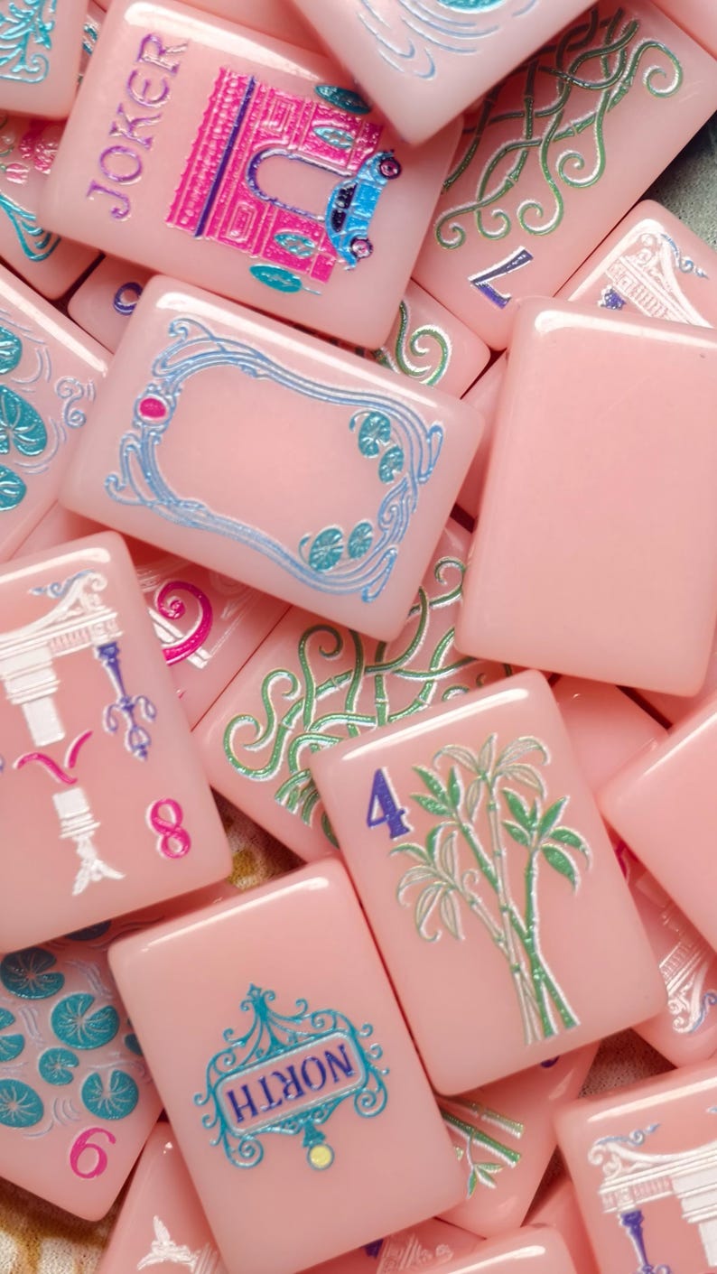 May include: A collection of rectangular mahjong tiles in a soft pink hue, featuring engraved designs in metallic blue, green, and silver. The tiles display various symbols, including the word JOKER, the word NORTH, a bamboo plant, and numbers.