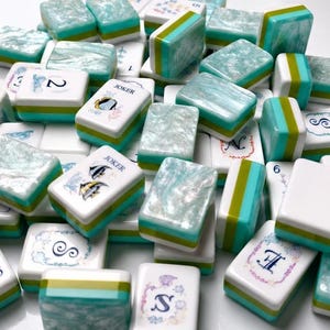 Op de afbeelding: Een verzameling rechthoekige mahjongtegels met een witte basis en een gelaagd ontwerp in de kleuren taling en limoengroen aan de zijkanten. De bovenkanten hebben een parelmoerachtige, gemarmerde talingafwerking of witte bedrukte vlakken.
