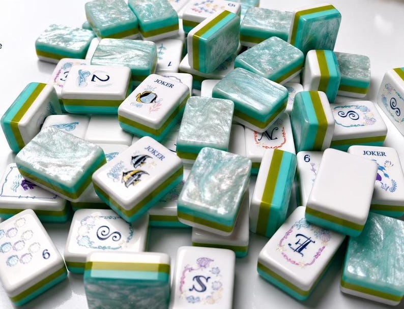 Op de afbeelding: Een verzameling rechthoekige mahjongtegels met een witte basis en een gelaagd ontwerp in de kleuren taling en limoengroen aan de zijkanten. De bovenkanten hebben een parelmoerachtige, gemarmerde talingafwerking of witte bedrukte vlakken.