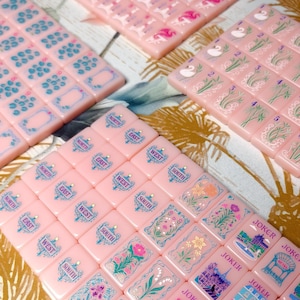 May include: A set of light pink mahjong tiles featuring various engraved designs including cardinal directions, floral patterns, swans, and joker cards with architectural illustrations. The tiles are arranged in rows and columns, displaying blue, green, and pink ink details on the smooth, rectangular surfaces.