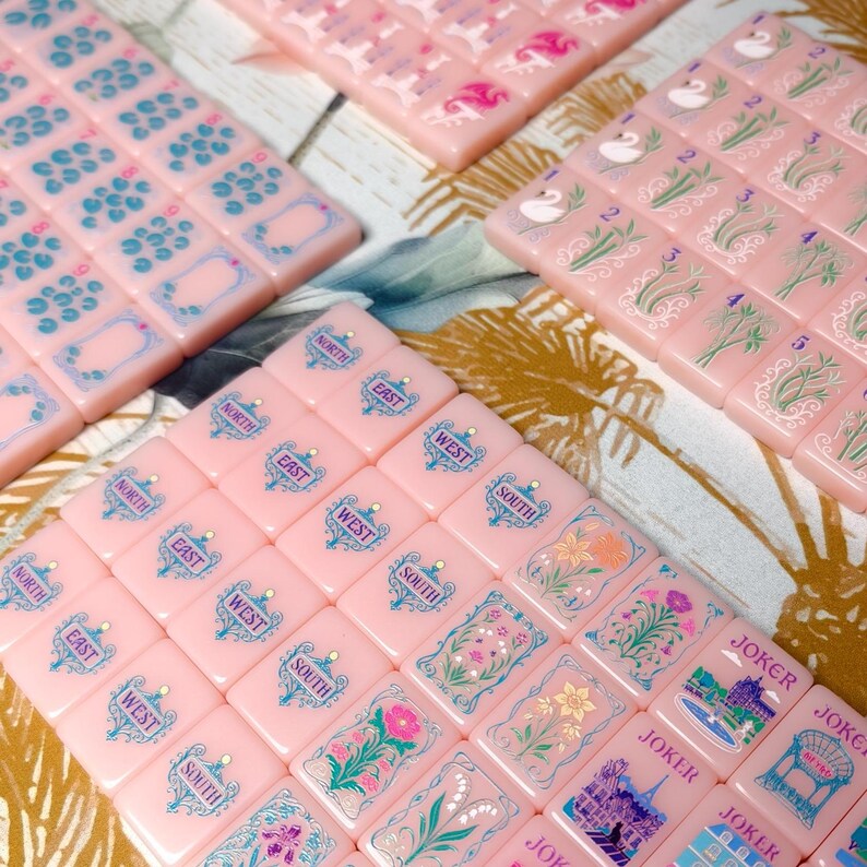 May include: A set of light pink mahjong tiles featuring various engraved designs including cardinal directions, floral patterns, swans, and joker cards with architectural illustrations. The tiles are arranged in rows and columns, displaying blue, green, and pink ink details on the smooth, rectangular surfaces.