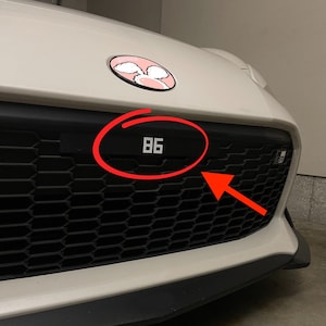 May include: A white car with a black grille and an "86" emblem. The car has a pink and white sticker on the bonnet.