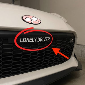 May include: White car with a black honeycomb grille. The grille has a black decal that reads "LONELY DRIVER" in white letters. The car has a pink and white decal on the hood.