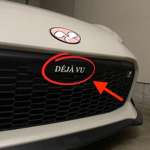 May include: A white car with a black grill and a silver emblem that says "DÉJÀ VU".