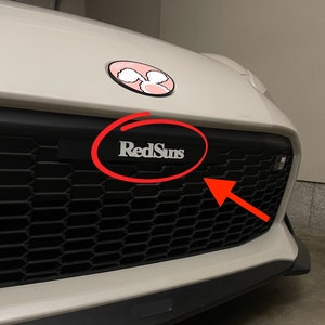 May include: A white car with a black honeycomb grille. The grille has a silver emblem that says "RedSuns".