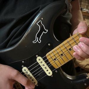 May include: Black electric guitar with a white outline of a dog on the body. The guitar has a light brown neck and white pickups. The strings are visible, and the guitar is being played.