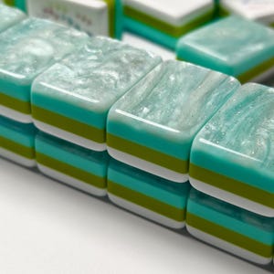 Op de afbeelding: Een set rechthoekige mahjongtegels gerangschikt in twee rijen. Elke tegel heeft een doorschijnende, parelmoerachtige lichtblauwe toplaag met een wervelende textuur, gevolgd door horizontale strepen van helder turquoise, limoengroen en wit aan de basis.