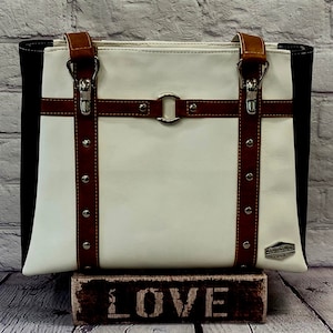 May include: A white and black tote bag with brown leather straps and buckles. The bag has a silver ring and a brown leather strap with stitching across the front. The bag is sitting on a wooden block with the word "LOVE" printed on it.