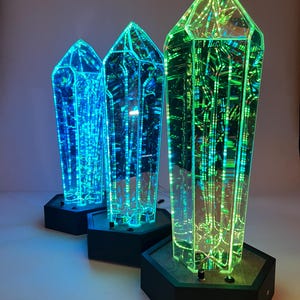 May include: Three illuminated crystal-shaped sculptures with a geometric design. The crystals are lit with blue and green lights, and sit on black hexagonal bases. The crystals have a reflective interior, creating a unique visual effect.