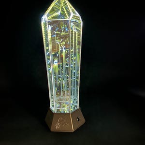 May include: A clear, multi-faceted crystal-shaped light fixture with an internal reflective structure that creates a glowing effect. The object is mounted on a hexagonal, metallic-toned base featuring an engraved fox head logo on the front side.