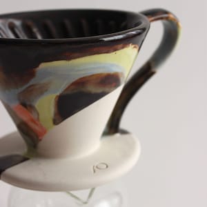 Peut inclure: Un verseur à café en céramique avec une base blanche et un intérieur coloré. L'intérieur présente des teintes marron, jaune, bleu et orange. La poignée et le bord sont brun foncé. La base a un petit motif estampé.