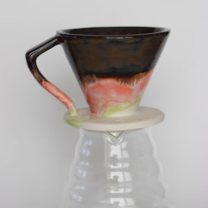 Könnte beinhalten: Ein Keramik-Pour-Over-Kaffeebereiter mit dunkelbraunem Oberteil, das in Korallen- und Grüntöne übergeht. Er hat einen Griff und sitzt auf einer klaren Glaskaraffe, ideal zum Kaffee kochen.