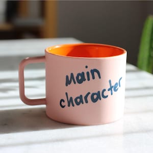 Könnte beinhalten: Eine rosa Keramiktasse mit orangefarbenem Innenraum. Die Tasse hat einen Henkel und die Worte "main character" sind in dunkelblauer Schrift auf die Seite geschrieben. Die Tasse steht auf einer weißen Oberfläche.