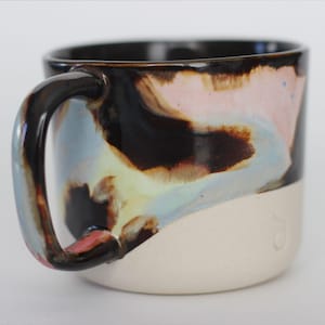 Könnte beinhalten: Eine Keramik-Tasse mit schwarzem Innenraum und weißem Boden. Der obere Teil hat ein farbenfrohes, wirbelndes Design in Rosa, Blau, Braun und Gelb. Der Griff ist schwarz und glänzend.