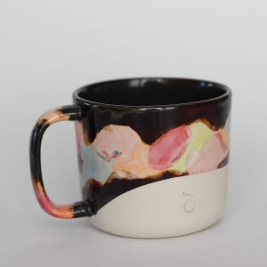 May include: A ceramic mug with a black interior and a white base. The upper portion features a colorful, abstract design with pink, yellow, and blue hues. The handle transitions from orange to pink. A small, stylized "jo" is on the base.