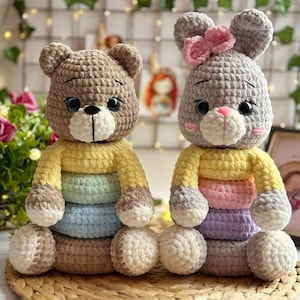 Può includere: Due animali di peluche all'uncinetto, un orso e un coniglio, entrambi con magliette a righe colorate. L'orso è marrone e il coniglio è grigio. Entrambi hanno grandi occhi neri e nasi bianchi.