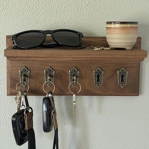 Puede incluir: Un estante de pared rectangular de madera con cinco ganchos de latón antiguo en forma de cerradura. Unas gafas de sol negras y una pequeña taza de cerámica con un degradado de color beige y marrón descansan sobre el borde superior.