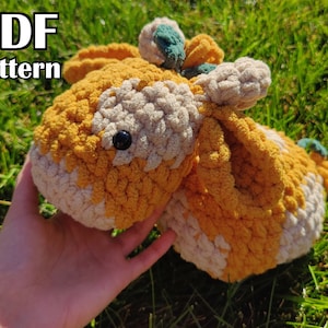 May include: A crocheted stuffed animal in the shape of a cow with a yellow and cream color pattern. The cow has a black eye and a green flower on its head. The cow is sitting on a bed of green grass.