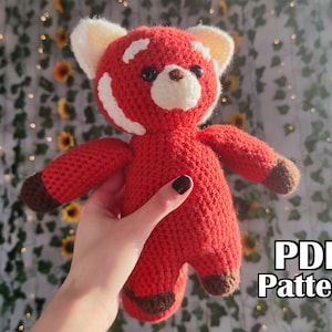May include: A crocheted red panda plush toy with white accents and brown details. The toy is being held by a hand in front of a green leafy background. The text "PDF Pattern" is visible in the lower right corner of the image.
