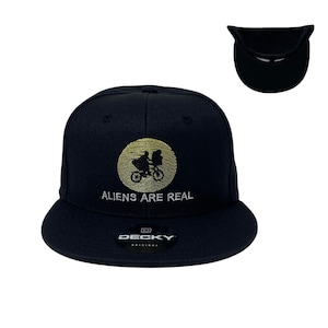 Peut inclure: Casquette de baseball noire avec une image brodée en or d'un vélo et de deux personnages. Le texte "ALIENS ARE REAL" est brodé en blanc sous l'image. La casquette a une étiquette noire avec le mot "DECKY" en blanc.