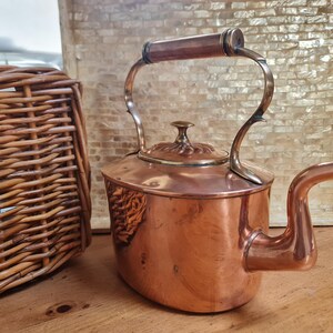 May include: A copper kettle with a wooden handle and a curved spout. The kettle has a lid with a decorative knob. The kettle is on a wooden surface. A wicker basket is visible on the left side of the image.