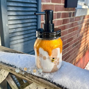 May include: A white ceramic soap dispenser with a black pump and a gold honey drip design. The dispenser has a bee and honeycomb pattern. The dispenser is sitting on a wooden surface covered in snow.