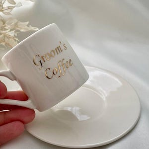May include: White ceramic coffee cup and saucer set. The cup has the words "Groom's Coffee" in gold lettering. The cup is held at an angle, with the saucer beneath it. The background is a white, silky fabric.