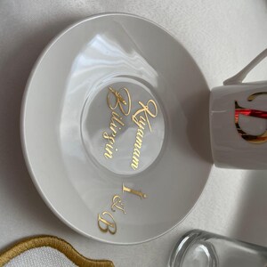May include: White ceramic teacup and saucer set with gold lettering. The saucer features the words "Bilgin Kayamam I & B" in gold script. The teacup has a gold design. A clear glass and a decorative coaster are also visible.