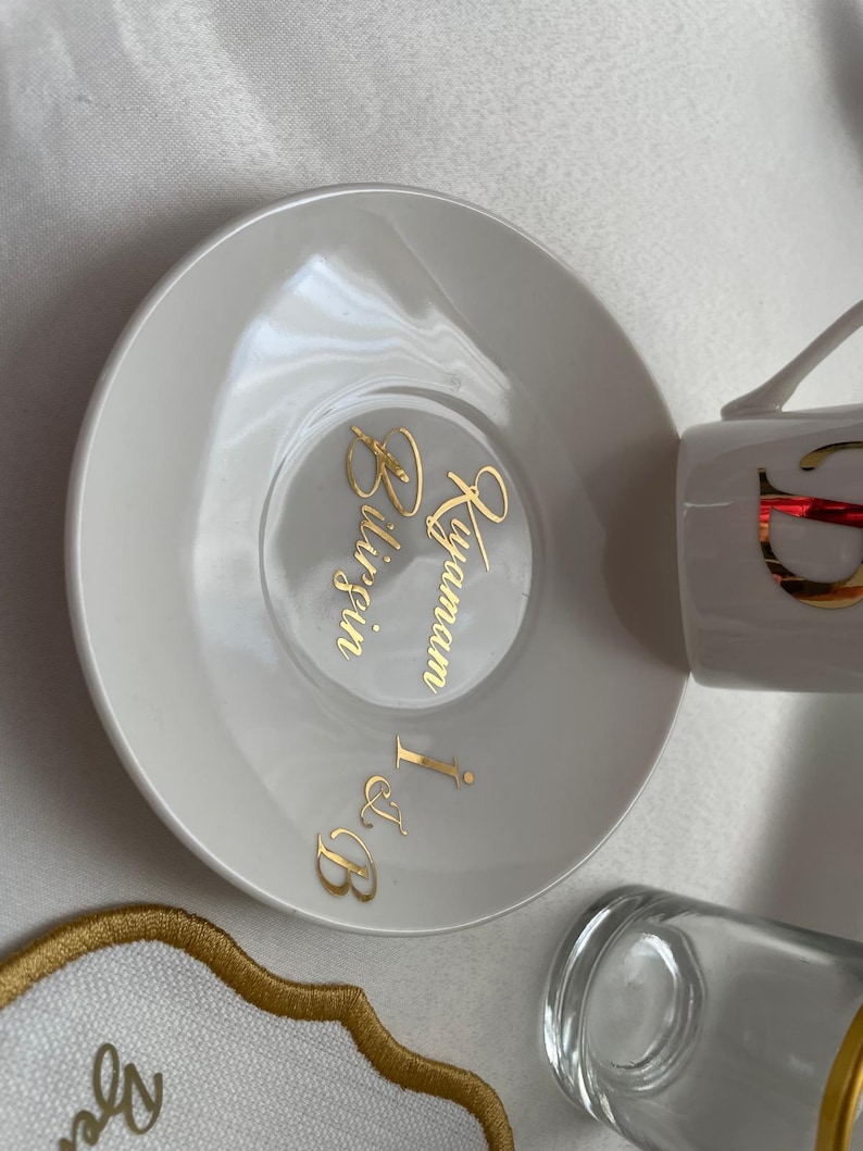 May include: White ceramic teacup and saucer set with gold lettering. The saucer features the words "Bilgin Kayamam I & B" in gold script. The teacup has a gold design. A clear glass and a decorative coaster are also visible.