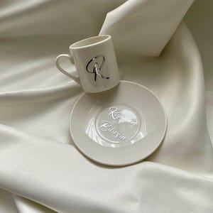 May include: A white ceramic coffee cup and saucer set. The cup has a silver letter "R" on the side. The saucer has the words "Kıyamam Bilirsin" in silver script. The set is on a white satin surface.