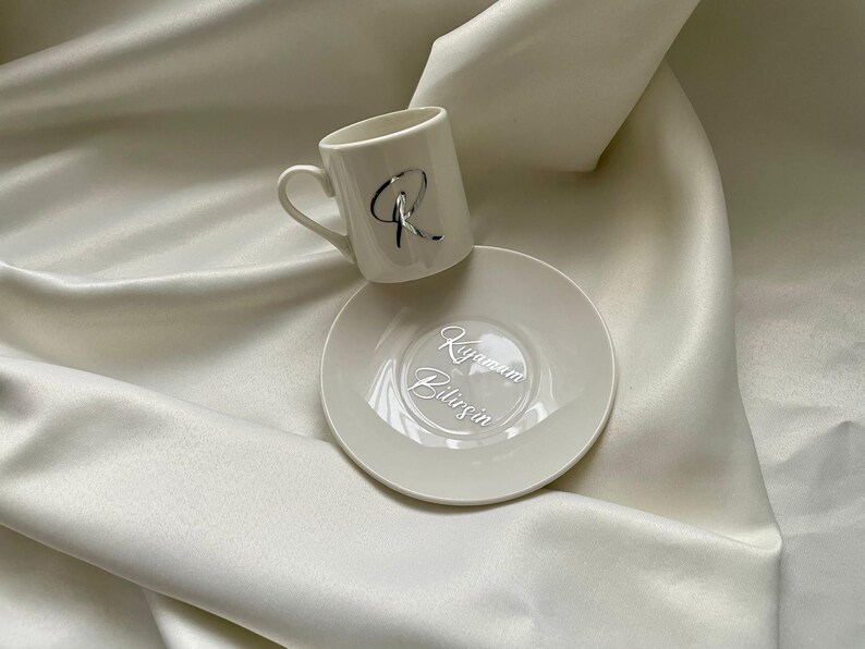 May include: A white ceramic coffee cup and saucer set. The cup has a silver letter "R" on the side. The saucer has the words "Kıyamam Bilirsin" in silver script. The set is on a white satin surface.