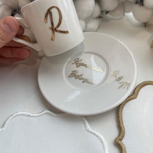 May include: White ceramic coffee cup and saucer set. The cup features a gold letter "R" and the saucer has gold script. The set is on a white surface with white and gold embroidered details.