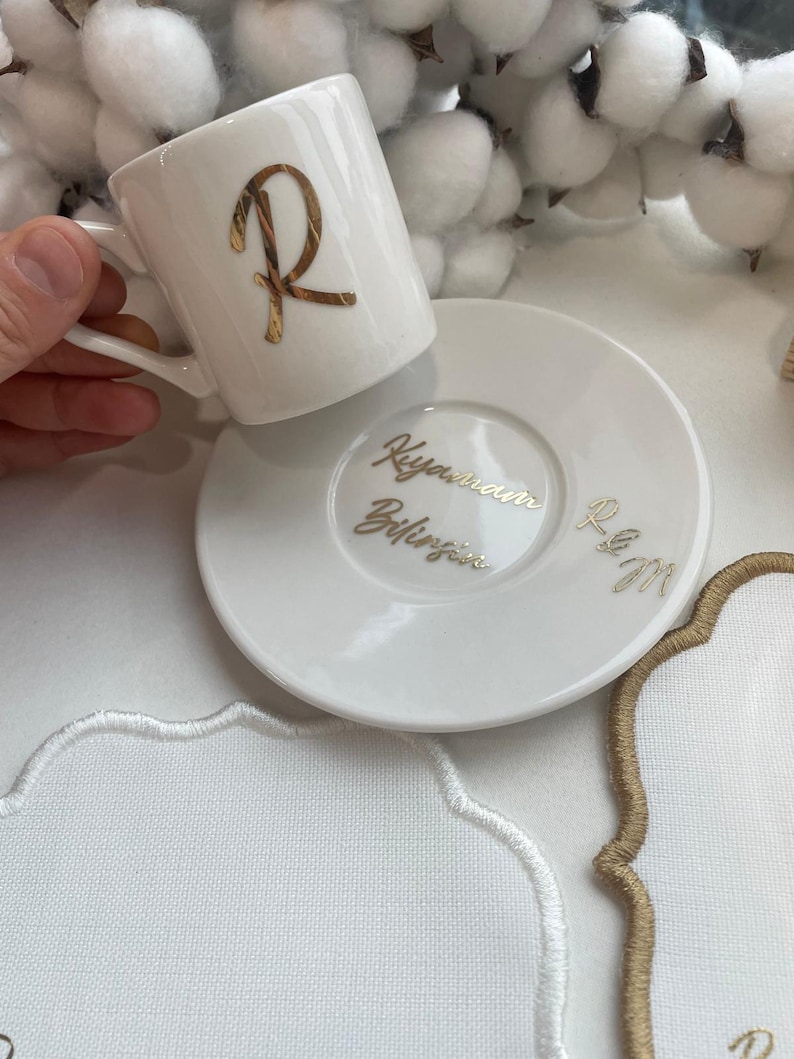 May include: White ceramic coffee cup and saucer set. The cup features a gold letter "R" and the saucer has gold script. The set is on a white surface with white and gold embroidered details.