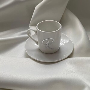 May include: A white ceramic espresso cup and saucer set. The cup features a handle and a raised, stylized letter "R" on the front. The set is placed on a white, textured fabric, creating a minimalist aesthetic.