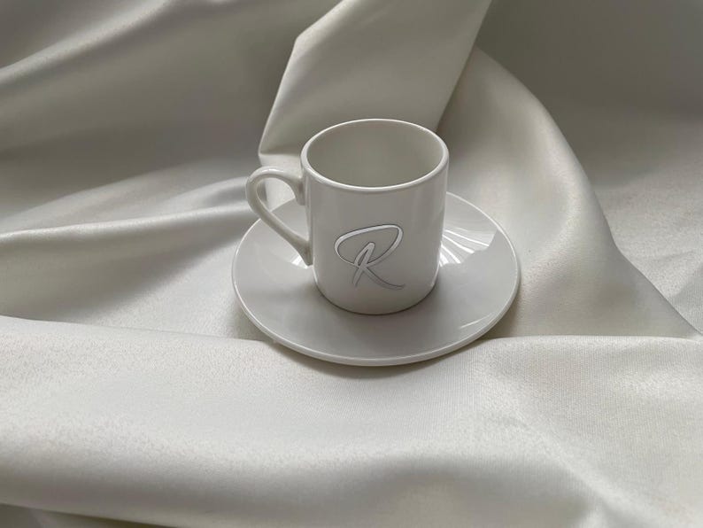 May include: A white ceramic espresso cup and saucer set. The cup features a handle and a raised, stylized letter "R" on the front. The set is placed on a white, textured fabric, creating a minimalist aesthetic.