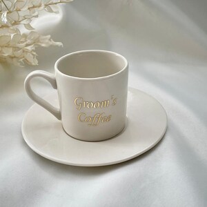 May include: A cream-colored ceramic coffee cup and saucer set. The cup has a handle and the words "Groom's Coffee" in gold lettering. The saucer is round and the same color as the cup. The set is on a white surface.
