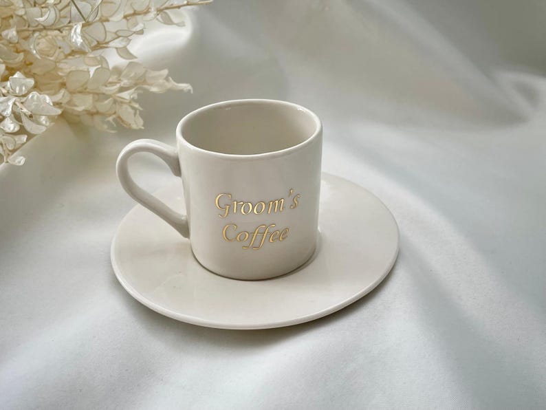 May include: A cream-colored ceramic coffee cup and saucer set. The cup has a handle and the words "Groom's Coffee" in gold lettering. The saucer is round and the same color as the cup. The set is on a white surface.