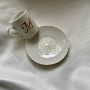 May include: White ceramic coffee cup and saucer set. The cup features a gold letter "M" and the saucer has gold text. The set is displayed on a white, silky surface. A simple and elegant design.