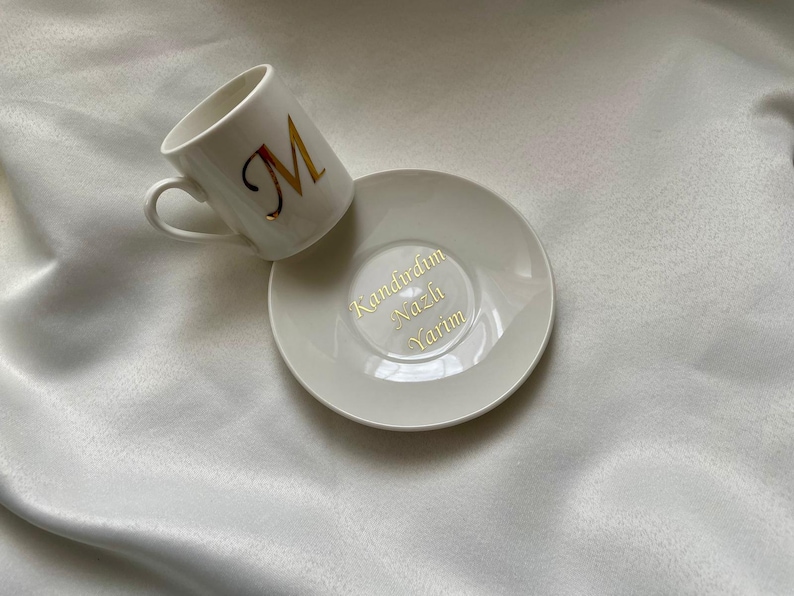 May include: White ceramic coffee cup and saucer set. The cup features a gold letter "M" and the saucer has gold text. The set is displayed on a white, silky surface. A simple and elegant design.