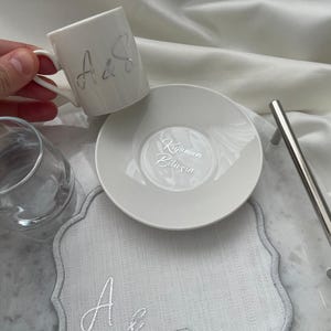 May include: A white ceramic coffee cup and saucer set with silver lettering. The cup has the letters "A&S" on the side. The saucer has text in a script font. A gray placemat with the letters "A&S" is also present.