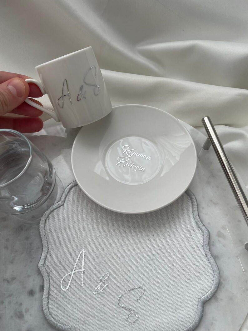 May include: A white ceramic coffee cup and saucer set with silver lettering. The cup has the letters "A&S" on the side. The saucer has text in a script font. A gray placemat with the letters "A&S" is also present.