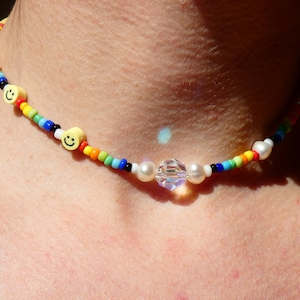 May include: A colorful beaded choker necklace with a crystal and pearl charm. The necklace features yellow smiley face beads and rainbow colored beads.