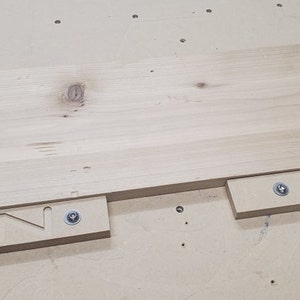 Op de afbeelding: Een close-up van een licht houten plank die wordt gezaagd met een mal. De mal heeft twee stukken hout met de letter "N" uitgesneden. De mal is met schroeven aan de plank bevestigd.