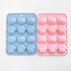 May include: Two silicone molds for making chocolate or candy. One mold is light blue and the other is pink. Both molds have a design of cat faces and paw prints.