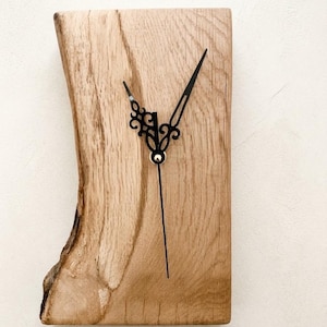 May include: A rectangular wall clock made from a single piece of light-coloured wood with a natural live edge on the left side. The clock features black metal hands with ornate, decorative cutouts and a simple, straight second hand.